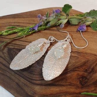 A pair of handmade silver sage-leaf and seaglass dangly earrings placed on some olive wood and with herb flowers