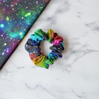 A cotton scrunchie with a starburst space pattern featuring vibrant bursts of colour resembling stars and galaxies against a dark background.