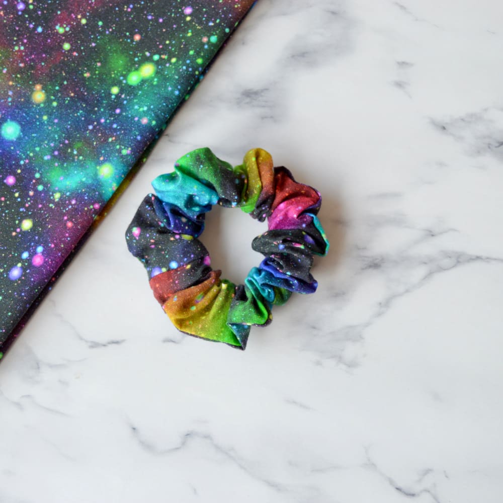 A cotton scrunchie with a starburst space pattern featuring vibrant bursts of colour resembling stars and galaxies against a dark background.