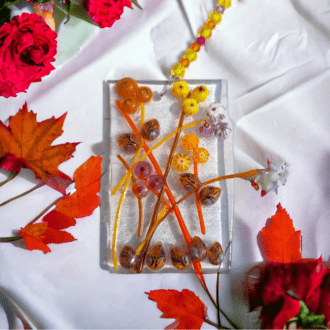 A close-up photo of a handmade fused glass suncatcher with an autumn theme. The square-shaped suncatcher is made of clear glass decorated with pieces of orange, yellow, and red coloured glass, creating an abstract autumn design. The suncatcher hangs from a jewellery wire with colourful beads and measures approximately 19.5 cm in total length. The fused glass piece itself measures 5 cm wide and 8 cm long.