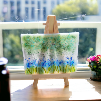 A close-up photo of a handmade fused glass artwork featuring blue and green coloured glass to depict a bluebell forest scene. The fused glass art measures 11 cm wide and 8 cm tall and sits on a mini wooden easel. The easel is painted brown and measures 15 cm high, 8 cm wide at the base, and 10 cm deep.