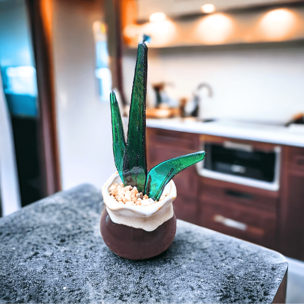 A close-up photo of a handmade fused glass snake plant in a ceramic pot. The snake plant is made of green glass and has a long, upright form with vertical stripes. The ceramic pot is white and measures 5 cm high and 6 cm wide. The fused glass snake plant is 11 cm tall and 8 cm at its widest.