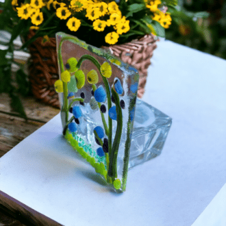 A close-up photo of a handmade fused glass tealight holder. The tealight holder is made of clear glass with a fused design of blue and green flowers on the front. It measures approximately 7.5 cm in height and width, and 6 cm deep. The tealight holder sits on a white background.
