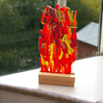 A photo of a glass candle holder on a wooden stand. The clear glass holder features a colourful fused glass design with red and orange glass on the front. It rests on a wooden base