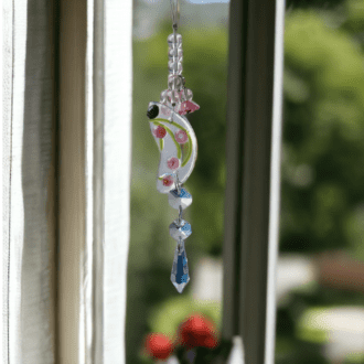 A close-up photo of a one-of-a-kind, handmade fused glass suncatcher in an organic shape. The piece appears to have been created from clear glass with a light purple and pink flowers. It is accented with a string of colourful beads and lucite flowers, and it hangs from a window hanger. The total length of the suncatcher is approximately 20 cm, with the glass piece itself measuring 6 cm long and 3 cm wide at its widest point.