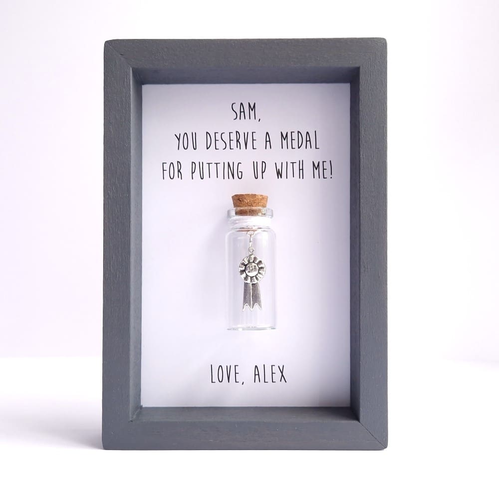 Small grey frame with a personalised quote and miniature glass bottle containing a medal