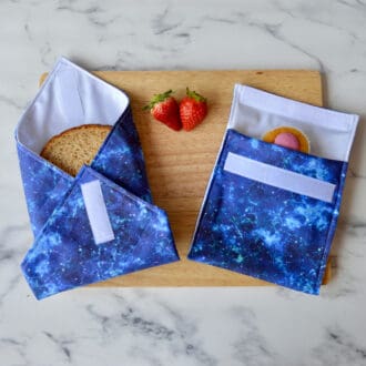 Blue cosmic sandwich wrap and snack bag on a wooden board. A sandwich is partially wrapped in the sandwich wrap, a cupcake is inside the snack bag, and fresh strawberries are placed on the board.