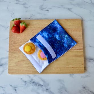 A blue cosmic reusable snack bag is displayed on a wooden board with cupcakes and strawberries. The bag features a pattern reminiscent of a star-filled night sky