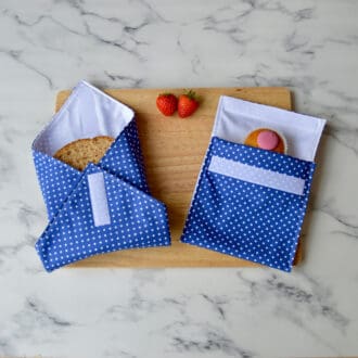 Sandwich wrap with a matching snack bag on a wooden board. Fabric is blue with white dots. A sandwich is partially wrapped in the sandwich wrap, a cupcake is inside the snack bag, and fresh strawberries are placed on the board.