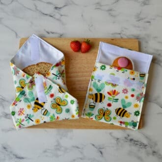 Cream cotton fabric sandwich wrap and snack bag with a pattern of bees, butterflies, caterpillars and flowers on a wooden board. A sandwich is partially wrapped in the sandwich wrap, a cupcake is inside the snack bag, and fresh strawberries are placed on the board.