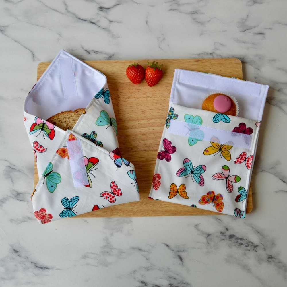 White sandwich wrap and snack bag with multi coloured butterflies on a wooden board. A sandwich is partially wrapped in the sandwich wrap, a cupcake is inside the snack bag, and fresh strawberries are placed on the board.