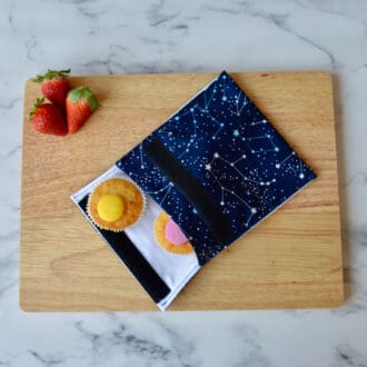A navy reusable snack bag with white constellations is placed on a wooden board alongside cupcakes and strawberries.
