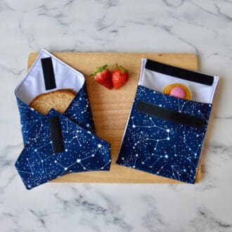Constellation-themed sandwich wrap and snack bag on a wooden board. The wrap and bag feature dark blue fabric with white star patterns, evoking a night sky filled with constellations. A sandwich is partially wrapped in the sandwich wrap, a cupcake is inside the snack bag, and fresh strawberries are placed on the board.