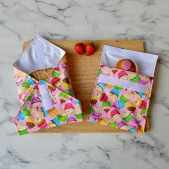 Pink cotton fabric sandwich wrap and snack bag with a cupcake pattern on a wooden board. A sandwich is partially wrapped in the sandwich wrap, a cupcake is inside the snack bag, and fresh strawberries are placed on the board.