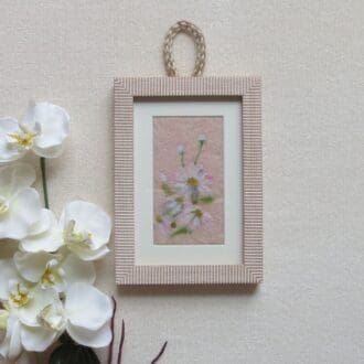 Handmade needle felted white daisies on a peach felt background in a pale wood effect frame with a jute cord for hanging.