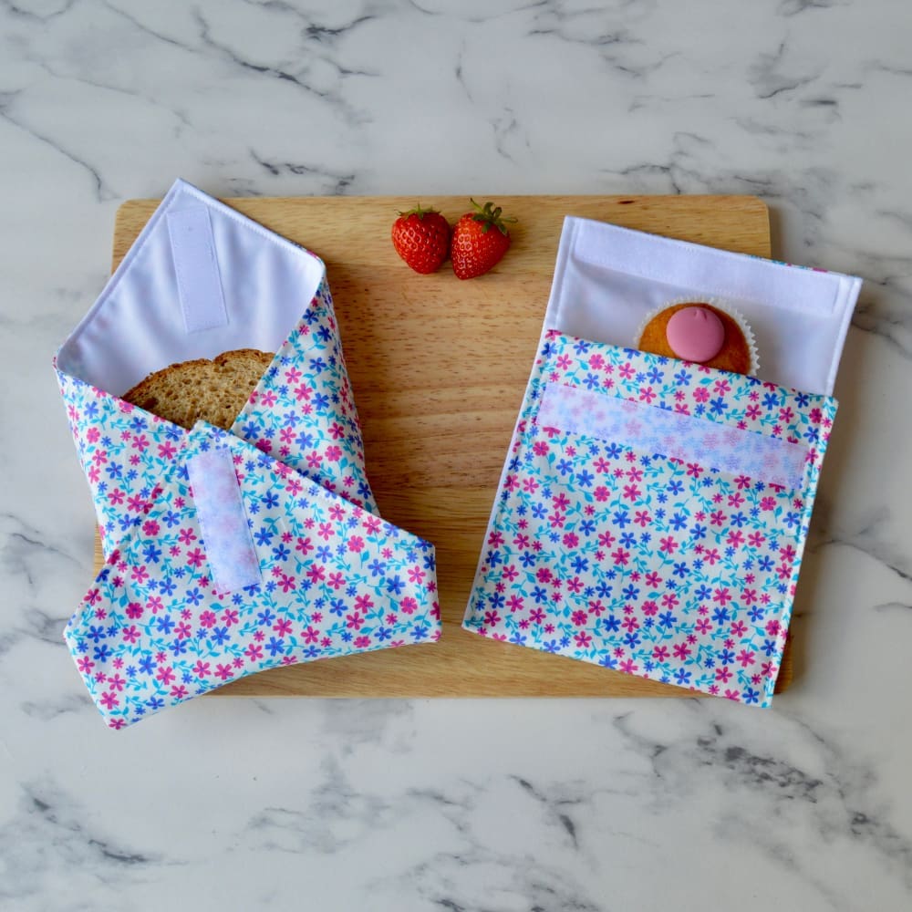 Pink and blue daisy sandwich wrap and snack bag on a wooden board. A sandwich is partially wrapped in the sandwich wrap, a cupcake is inside the snack bag, and fresh strawberries are placed on the board.