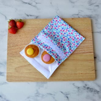 A floral pink and blue daisy snack bag is displayed on a wooden board with cupcakes and strawberries.