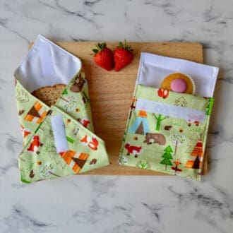 Green Forest Animals snack bag and matching sandwich wrap on a wooden board. Fabric has a green background with trees, foxes, bears, hedgehogs and owls. A sandwich is partially wrapped in the sandwich wrap, a cupcake is inside the snack bag, and fresh strawberries are placed on the board.