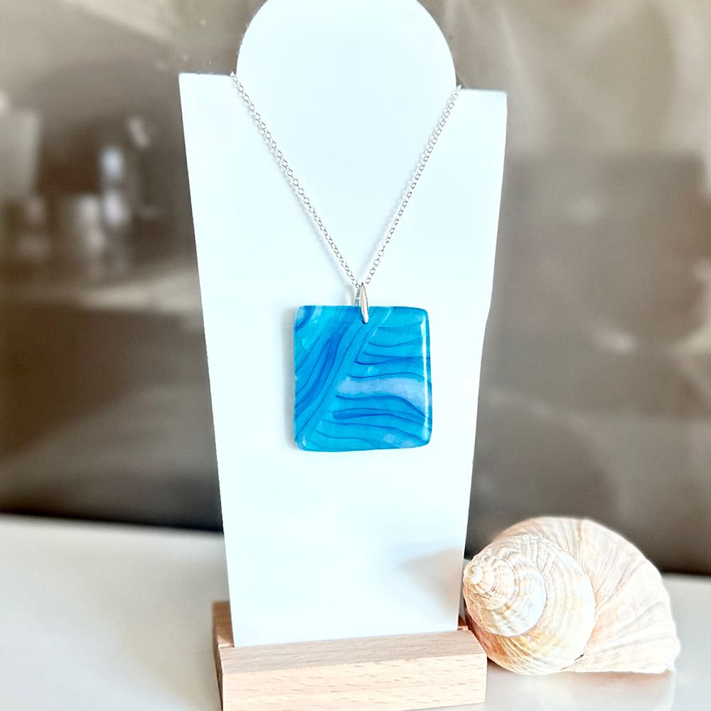 A square turquoise blue polymer clay pendant with a sterling silver chain on a white display stand.