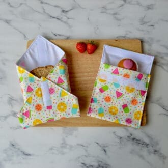 Matching sandwich wrap and snack bag on a wooden board. Fabric is pale yellow with ice cream and fruit. A sandwich is partially wrapped in the sandwich wrap, a cupcake is inside the snack bag, and fresh strawberries are placed on the board.