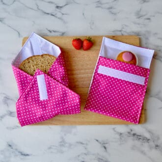 Sandwich wrap and matching snack bag on a wooden board. Fabric is pink with white spots. A sandwich is partially wrapped in the sandwich wrap, a cupcake is inside the snack bag, and fresh strawberries are placed on the board.