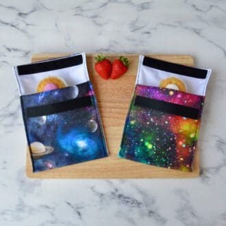 Two space-themed snack bags on a wooden board. One features planets on a dark background, and the other displays multi-coloured clusters of stars on a dark background. Cupcakes and fresh strawberries are also on the board.