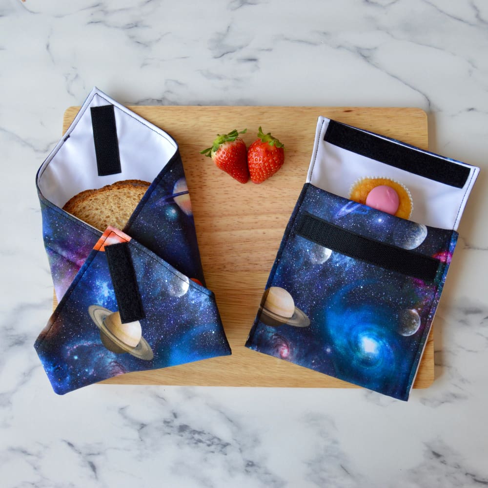 Planets space-themed sandwich wrap and snack bag on a wooden board. The fabric is navy with planets and star clusters. A sandwich is partially wrapped in the sandwich wrap, a cupcake is inside the snack bag, and fresh strawberries are placed on the board.
