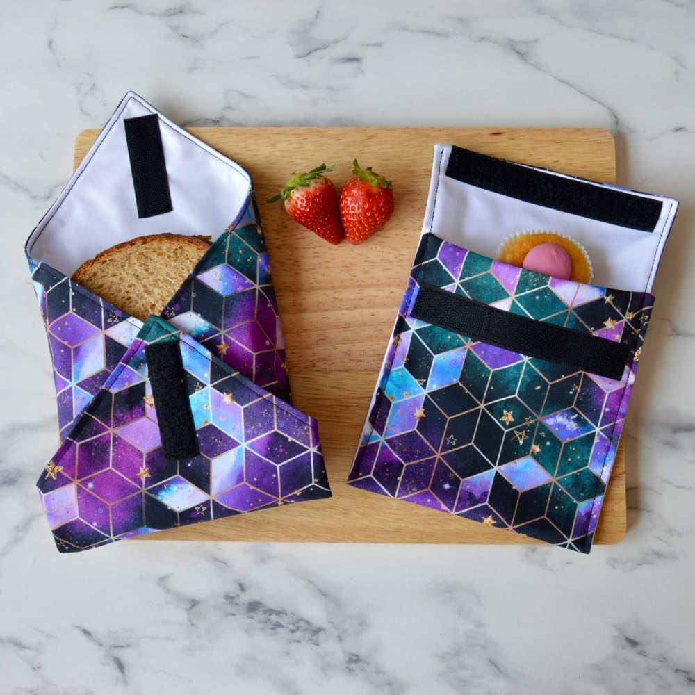 Purple, black and blue geometric fabric sandwich wrap and snack bag on a wooden board. A sandwich is partially wrapped in the sandwich wrap, a cupcake is inside the snack bag, and fresh strawberries are placed on the board.