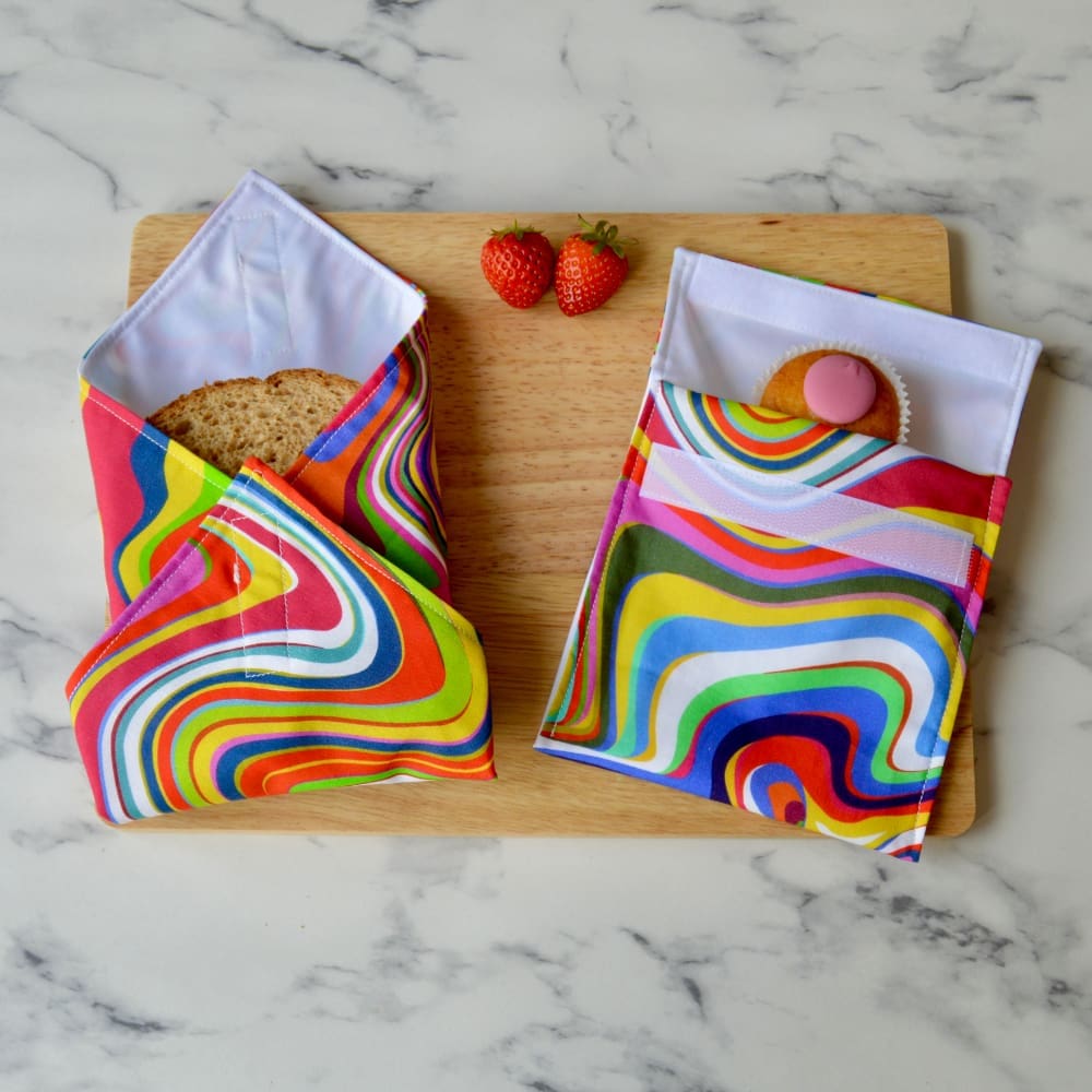 Retro rainbow multi coloured sandwich wrap and snack bag on a wooden board. A sandwich is partially wrapped in the sandwich wrap, a cupcake is inside the snack bag, and fresh strawberries are placed on the board.