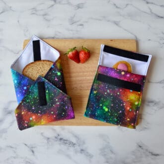 Starburst space-themed sandwich wrap and snack bag on a wooden board. A sandwich is partially wrapped in the sandwich wrap, a cupcake is inside the snack bag, and fresh strawberries are placed on the board.