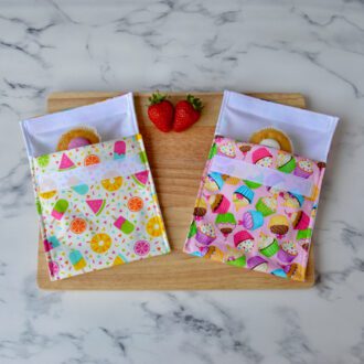 Two snack bags. one light yellow with ice creams and fruit and the other pink with cupcakes. Both laying on a wooden board with cupcakes inside and fresh strawberries placed around them