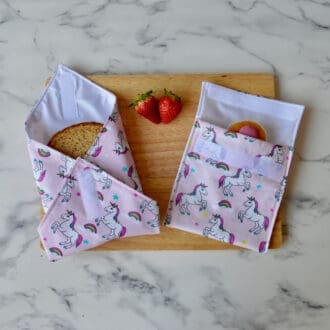 Light pink unicorn sandwich wrap and snack bag on a wooden board. A sandwich is partially wrapped in the sandwich wrap, a cupcake is inside the snack bag, and fresh strawberries are placed on the board.