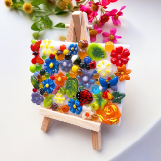 A vibrant fused glass 3D picture depicting a colourful meadow scene with intricate flowers, leaves, a ladybird, and a butterfly, all set on a mini wooden easel. The artwork features various bright colours and detailed textures, bringing the beauty of a garden indoors.