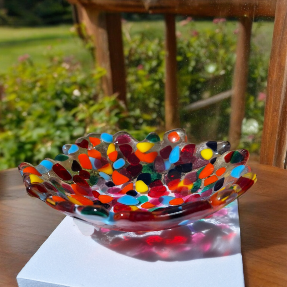 This image shows a handmade fused glass bowl featuring a vibrant multicoloured design. The bowl has an irregular, wavy edge and is adorned with a mosaic of colourful glass pieces in shades of red, orange, yellow, blue, green, and purple. The dynamic, playful pattern makes it a striking decorative piece, perfect for adding a pop of colour to any room. The bowl has a rounded bottom and is displayed against a white background.