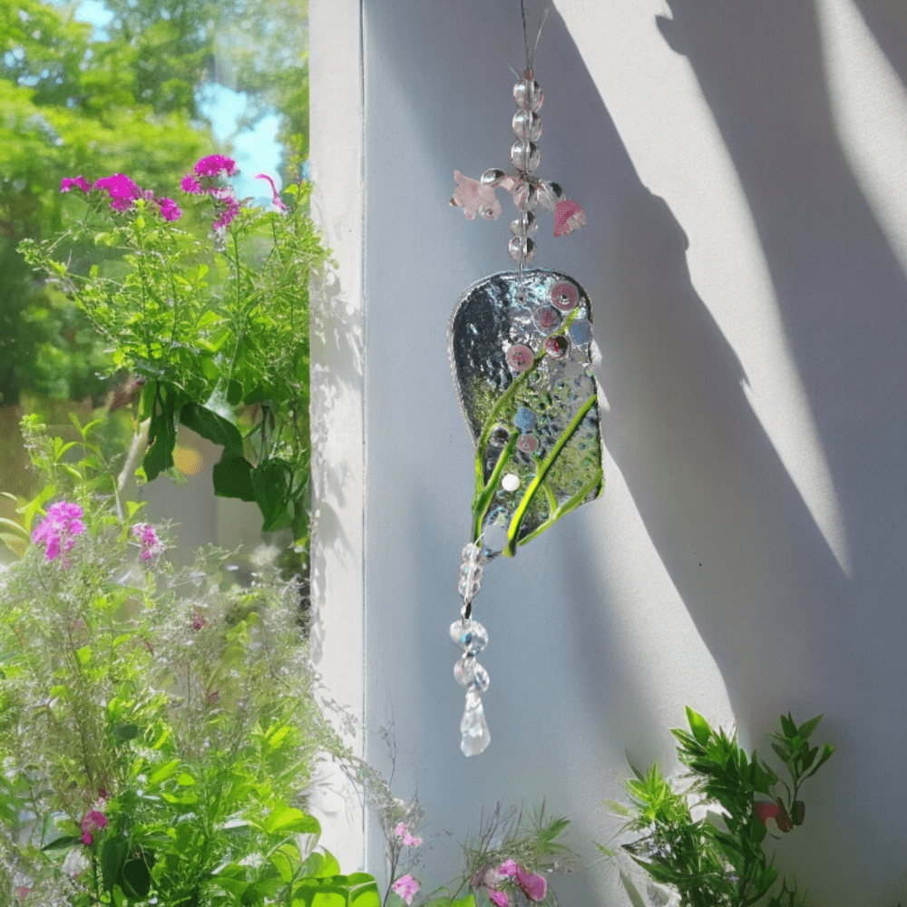 A fused glass suncatcher with a mix of transparent and green glass, adorned with small pink and white beads, hanging by a string with decorative elements. It casts a colourful light and shadow pattern on the wall beside a window, with vibrant green plants and pink flowers visible outside.