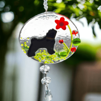 A photo of a handmade fused glass suncatcher featuring a black pug design. The suncatcher depicts a black pug sitting amongst colourful flowers in various shapes and sizes.