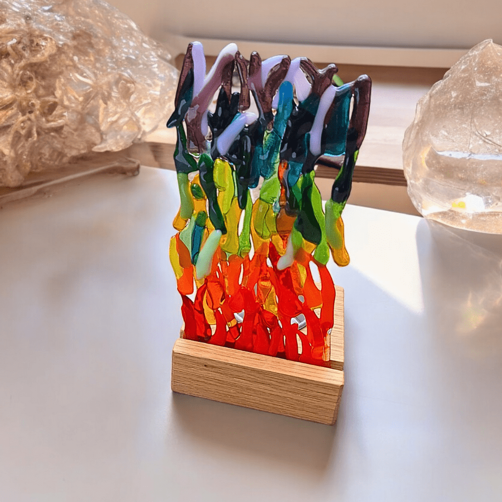 A vibrant fused glass sculpture featuring a mix of rainbow colours, set on a wooden stand. The glass pieces are arranged vertically, resembling abstract flames or waves, creating a dynamic and colourful display.