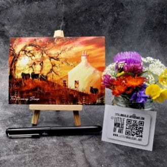 Image showing an A6 size greetings card, blank inside, on a small easel depicting an evening scene with guests beside the old croft house.