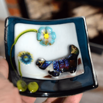 The image shows a handmade glass trinket dish. The dish features a colourful, iridescent design of a cat and two flowers. The cat is depicted in a shimmering, multi-coloured glass with a raised texture, and the flowers are rendered in vibrant blue and yellow hues. The dish itself is square with slightly curved edges and has a dark blue border around the rim. The overall design is artistic and whimsical, perfect for holding small items like jewellery or keys.