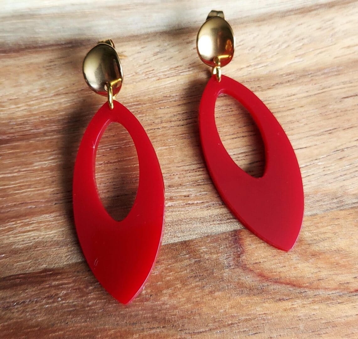 Mid century style oval drop hoop earrings handmade in berry red resin and hung from gold tone oval curved stud tops. Displayed on a wooden backdrop.