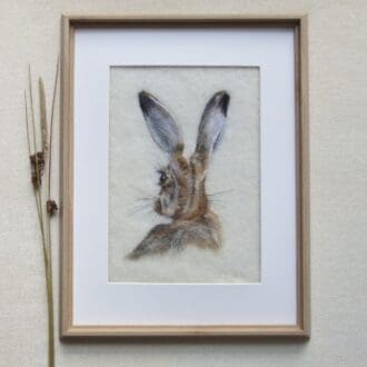 needle felted wool picture of a brown hare from behind showing the head, ears and shoulders. The head is turned to the left slightly so one eye is visible. The background is cream wool felt and the frame is a pale wood effect.