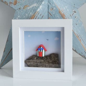 Miniature clay beach hut set on driftwood and framed in a white square frame.