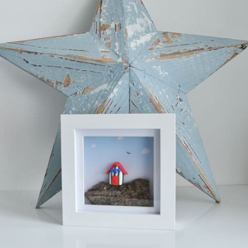 Miniature clay beach hut set on driftwood and framed in a white square frame.