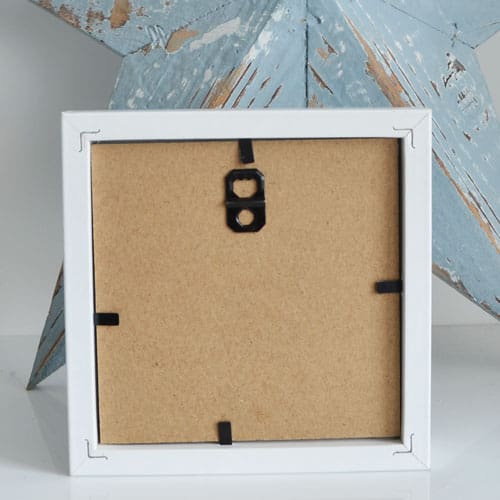 Miniature clay beach hut set on driftwood and framed in a white square frame.