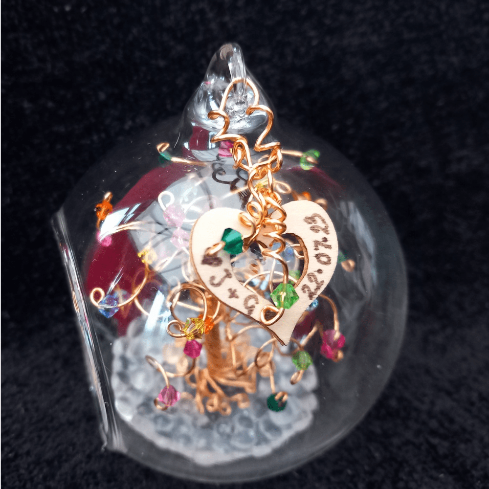 8cm glass bauble with a wooden heart charm wired onto the outside of it. The heart charm has a name and date written on it and decorated with gold wire and peridot and emerald crystal drops on spiral wire.