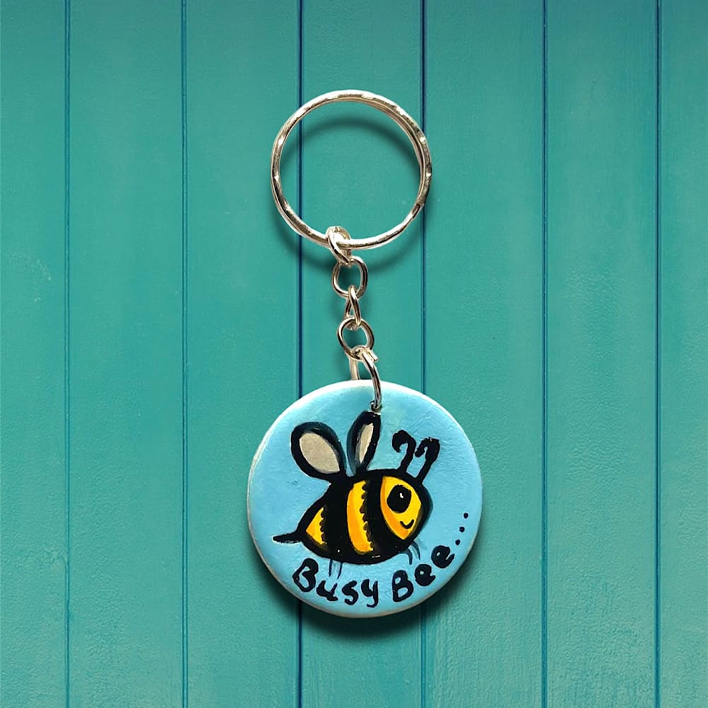 Handmade bumblebee keyring. Circular shaped keyring featuring a flying bumblebee on a blue background with the words "busy bee". Silver chain.