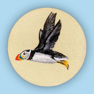 Handmade wood slice decoration featuring a puffin flying. Sage green background. 10 cm diameter.
