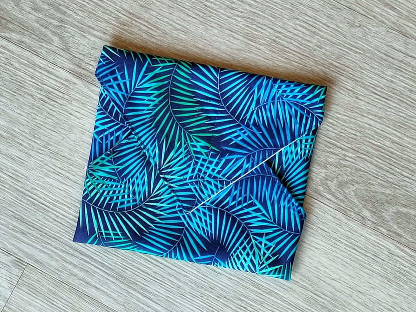 Reusable sandwich wrap approximately 30cm square, with a green and blue fern print cotton outer and food safe PUL lining.