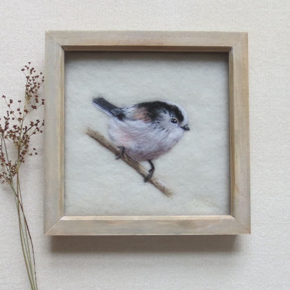 A handmade needle felted wool picture of a long tailed tit bird sat on a branch and looking to the right. The background is cream wool and the picture is in a square natural wood box frame.