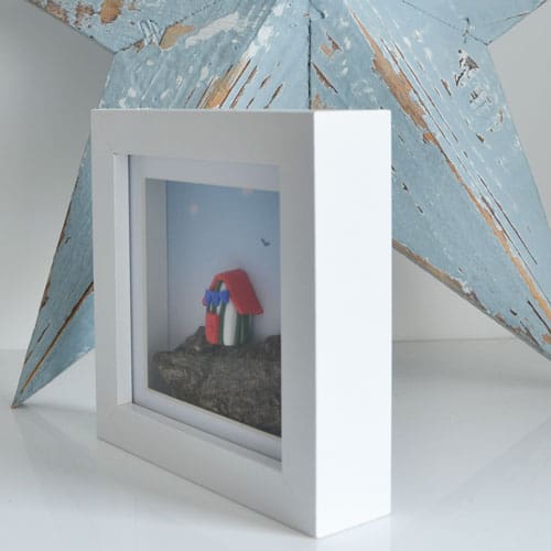 Miniature clay beach hut set on driftwood and framed in a white square frame.